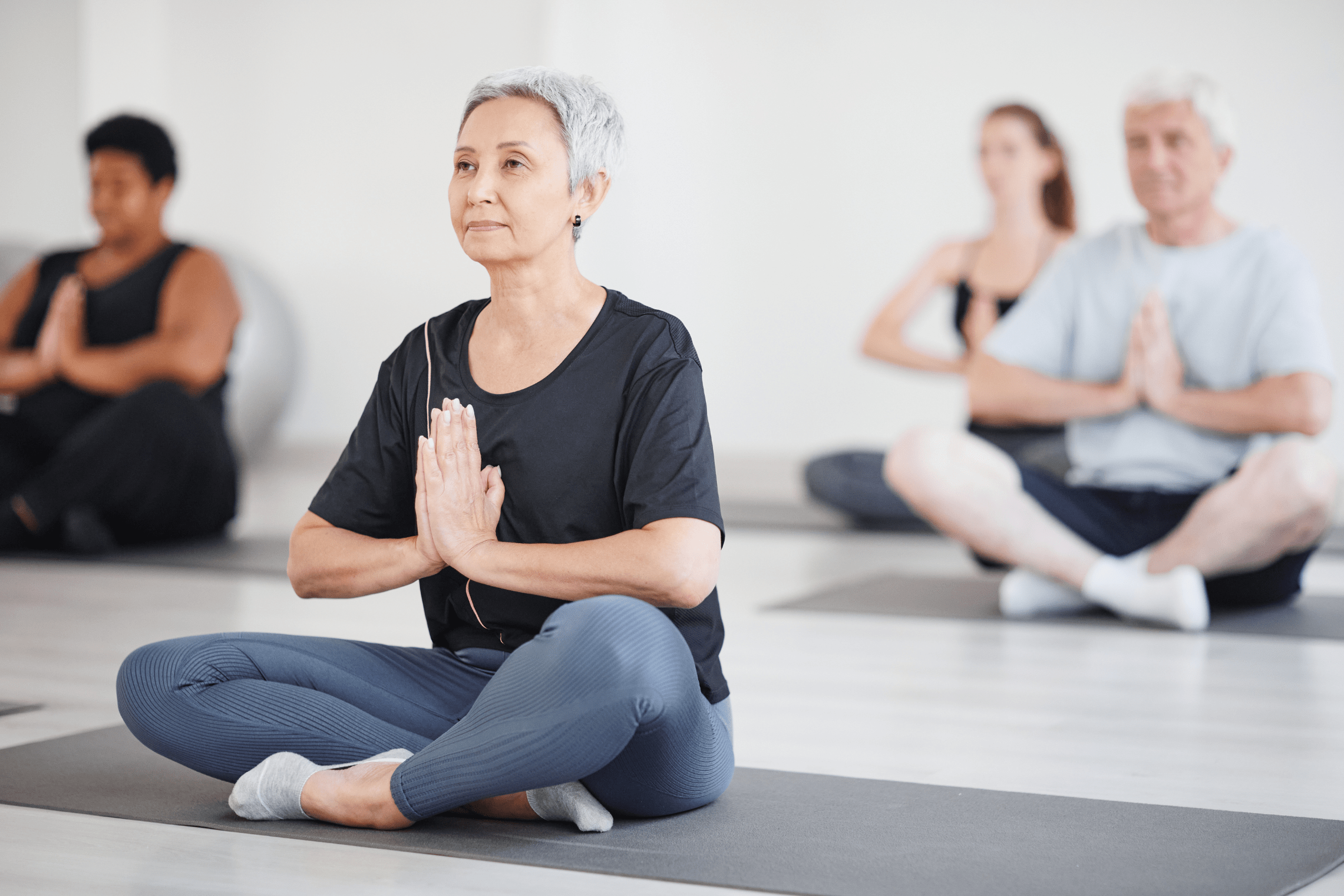 An image of a white haired woman sitting on a yoga mat in a class.